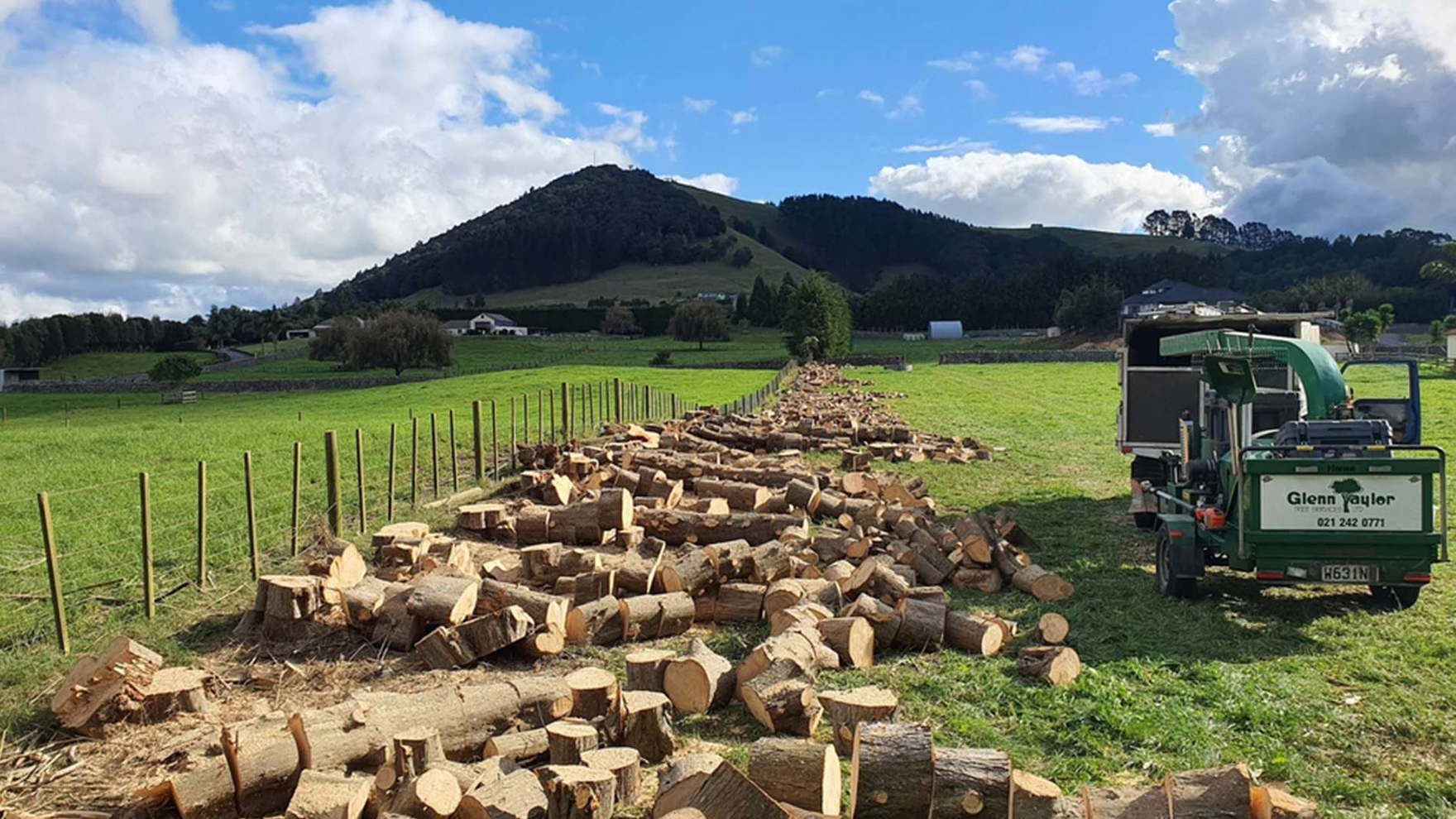 Rows of cut tree rounds along a farm fence line with the Glenn Taylor chipper parked on the grass and a hill behind