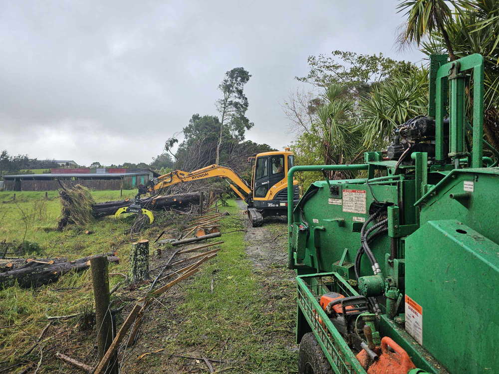 Excavator stacking felled trees along a rural fence line with the wood chipper in the foreground